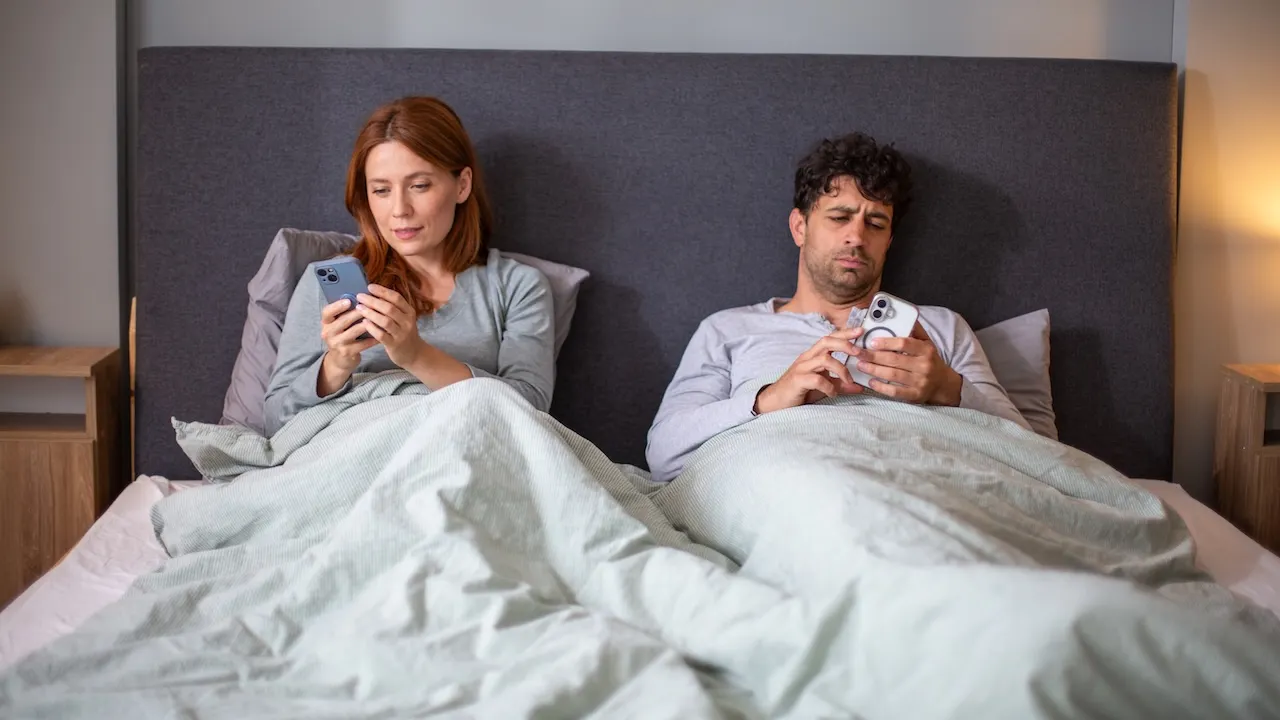 Pasangan suami istri duduk berdampingan di tempat tidur sambil menatap layar ponsel masing-masing